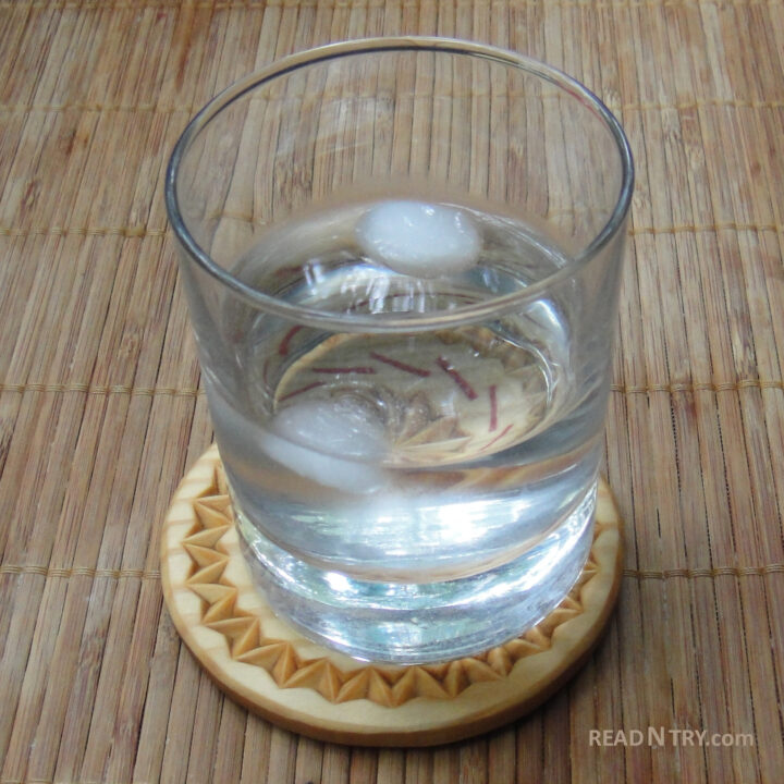 A glass with water on a chip carved coaster. The coaster chip carving decoration is refracted but visible through the glass.