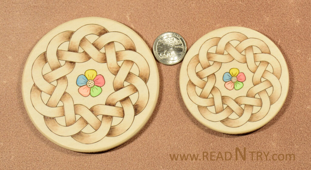 Two wooden coasters with a round Celtic knot pattern burned on the surface. The center of the coaster is decorated with a colorful flower. The image includes a quarter for scale.