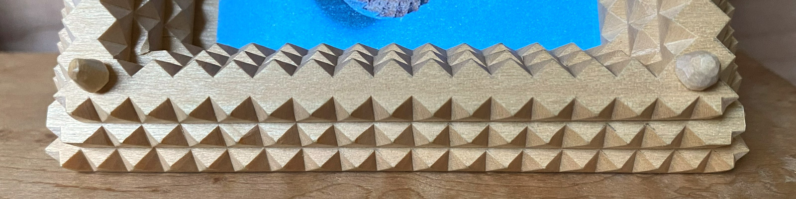 A close-up of a wooden picture frame carved in the Tramp Art style from basswood. The frame consists of three layers with carved notches.