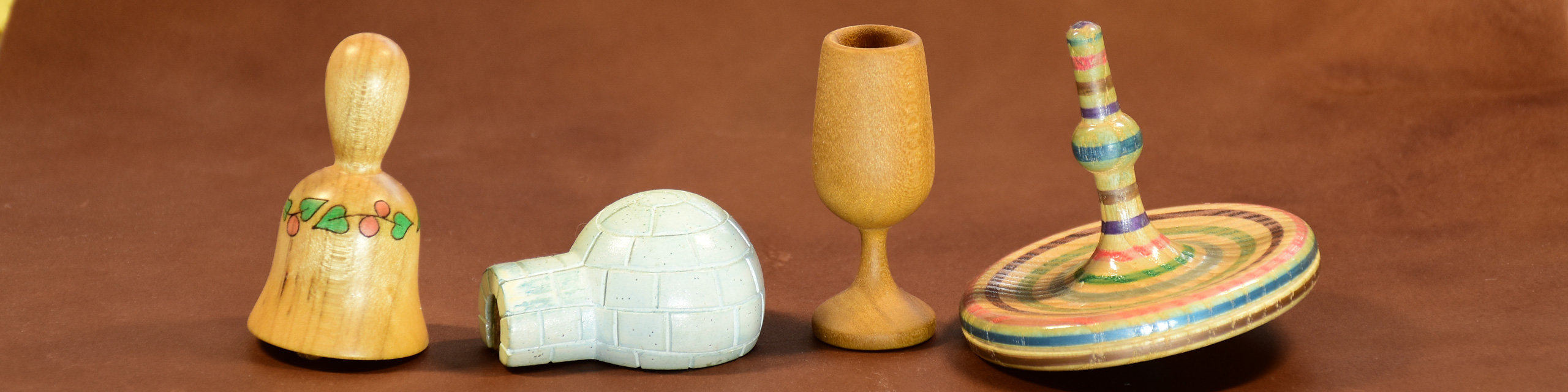 Four miniature turned pieces, each under 2.5 inches tall: a maple bell, a bass wood igloo with carving, a poplar goblet, and a sycamore spinning top.