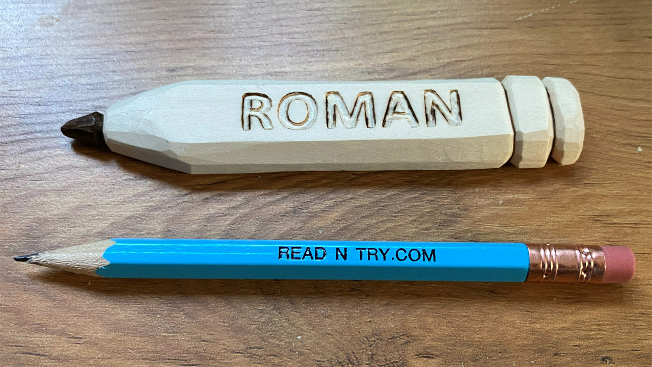 Carved and partially woodburned pencil-shaped name badge, almost ready for painting. It is shown alongside a real pencil. The pencil-shaped whittling was done by using the READ N TRY woodcarving pattern generator.